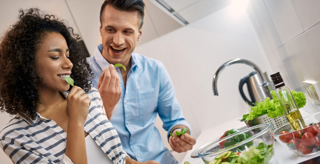 A white man and an african american woman stand in a kitchen eating green vegetables. They smile at each other.