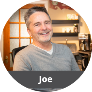 In a circular frame: An older white male with short gray hair sits in an office chair. He wears a gray sweater and is smiling. Across the bottom is a dark gray banner with white text in it that reads "Joe".
