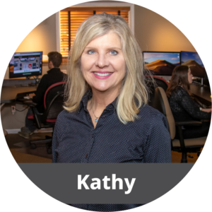 In a circular frame: An older white female with blonde hair stands in an office. She wears a navy blue button down with white dots on it and smiles. Across the bottom is a dark gray banner with white text in it that reads "Kathy".