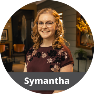 In a circular frame: A young adult white female with blonde hair and glasses stands in an office. She wears a red blouse with flowers on it and smiles. Across the bottom is a dark gray banner with white text in it that reads "Symantha".